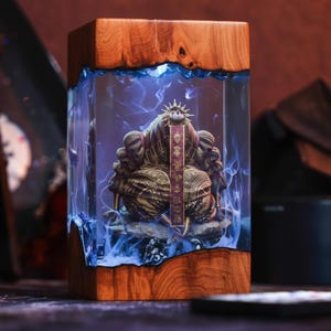 May include: A wooden and resin sculpture featuring a detailed golden figure within a clear resin block. The resin has a blue, smoky effect. The wooden base and top have a natural wood grain. A red banner with gold text is visible.