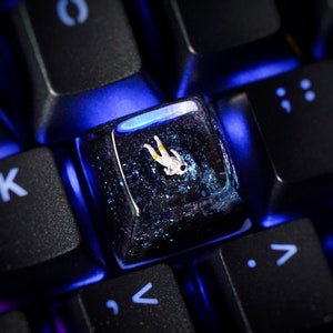 Pode incluir: Um close-up de um teclado de computador com uma única tecla substituída por uma tecla de resina transparente. A tecla contém uma pequena figura de astronauta dentro. O teclado é iluminado com retroiluminação azul.