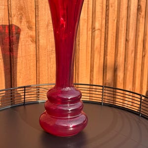 May include: A striking ruby red glass vase, featuring a flared top and a distinctive bulbous base. The vase has a long, slender neck and a series of rounded tiers. It is presented on a black metal tray, set against a wooden backdrop.