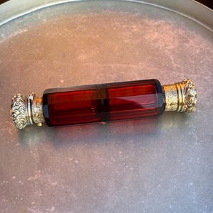 May include: A vintage perfume bottle with a rectangular, ruby-red glass body and ornate, gold-coloured metal caps. The caps feature intricate floral designs. The bottle is displayed on a silver-toned tray.