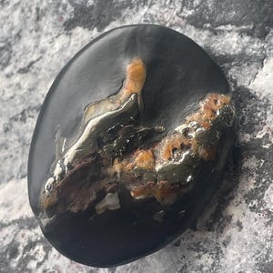 Puede incluir: Una piedra pulida de forma ovalada, con una base oscura, casi negra. La piedra presenta un llamativo patrón de depósitos minerales beige, marrón y blanco en un borde. La superficie es lisa y reflectante, mostrando variaciones naturales.