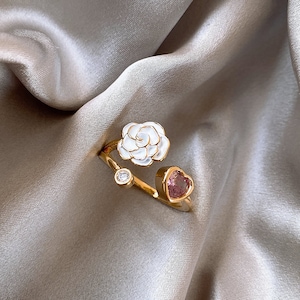 Könnte beinhalten: Ein goldener Ring mit einer weißen Blume und einem rosafarbenen herzförmigen Stein. Der Ring liegt auf einem hellbraunen Stoffhintergrund.