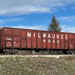 May include: A rusty red boxcar with white lettering that reads "MILWAUKEE ROAD" and "VMMX 61 47". The boxcar is on a set of train tracks.