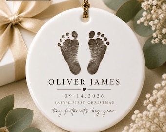 Op de afbeelding: Een rond wit keramieken ornament met donkerbruine babyvoetafdrukken bovenaan. Onder de afdrukken staat de tekst OLIVER JAMES, 09.14.2026, BABY'S FIRST CHRISTMAS en tiny footprints big year. Het hangt aan een goudkleurig koord.