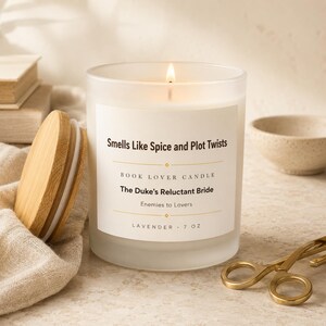 May include: A frosted glass candle with a wooden lid, featuring a white label that reads Smells Like Spice and Plot Twists, Book Lover Candle, The Duke's Reluctant Bride, Enemies to Lovers, Lavender, 7 oz. The candle has a lit wick.