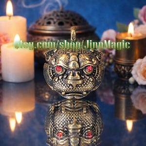 Puede incluir: Un quemador de incienso de latón de estilo antiguo, con forma de cabeza de animal estilizada con ojos de piedras preciosas rojas. La imagen incluye velas encendidas, flores y el texto "etsy.com/shop/JinnMagic".