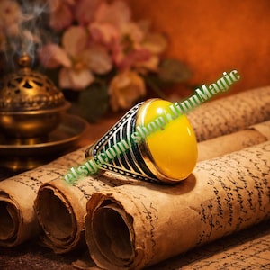 May include: A silver ring featuring a large, oval, yellow gemstone. The ring has a detailed black and silver band. The ring is displayed on a scroll of aged paper, with a golden incense burner and flowers in the background.