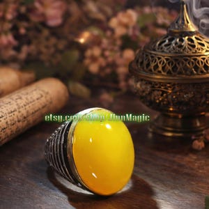 May include: A silver ring with a large, oval, yellow gemstone. The ring has a detailed, ribbed band. The image also shows a decorative incense burner, a scroll, and the text "etsy.com/shop/JinnMagic".