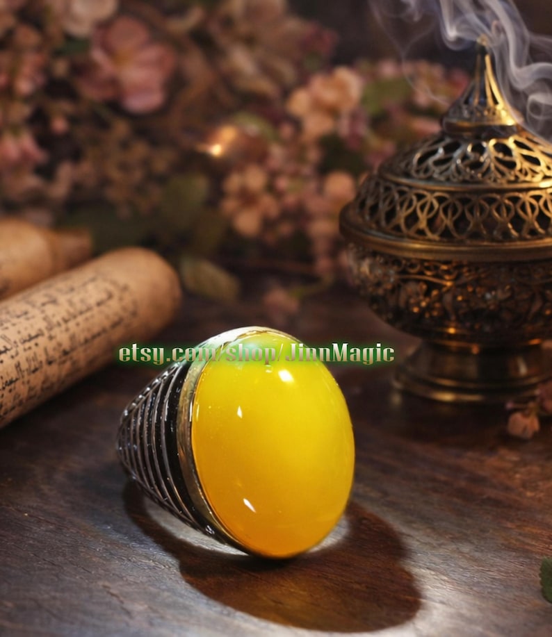 May include: A silver ring with a large, oval, yellow gemstone. The ring has a detailed, ribbed band. The image also shows a decorative incense burner, a scroll, and the text "etsy.com/shop/JinnMagic".