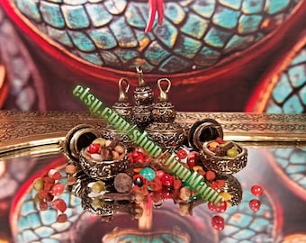 May include: Ornate, antique-style containers with lids, filled with colorful beads and small objects. The containers are gold-toned and sit on a reflective surface. The background features a decorative pattern with teal and red hues. The text "etsy.com/shop/InnMagic" is visible.