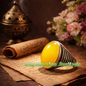May include: A silver ring with a large, oval, yellow gemstone. The ring is set against a backdrop of aged paper, a rolled scroll, and a decorative metal incense burner. The Etsy shop name "JinnMagic" is visible on the paper.