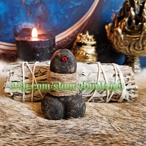 Può includere: Una scultura in pietra scura scolpita con un accento rosso, avvolta con spago. Due fasci di salvia bianca sono dietro la scultura. Una candela nera accesa e oggetti decorativi color oro sono sullo sfondo. Il nome del negozio Etsy è visibile.