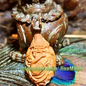 May include: A detailed, carved wooden figurine of a seated lion-like creature with an open mouth, set against a blurred background. A smaller, intricately carved wooden figure is in front. The background includes peacock feathers and the text "etsy.com/shop/JinnMagic".