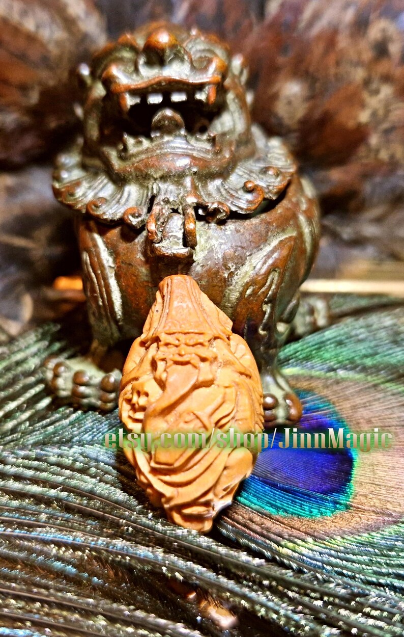 May include: A detailed, carved wooden figurine of a seated lion-like creature with an open mouth, set against a blurred background. A smaller, intricately carved wooden figure is in front. The background includes peacock feathers and the text "etsy.com/shop/JinnMagic".
