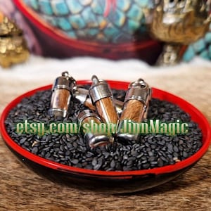 Puede incluir: Varios viales cilíndricos pequeños con tapas y bases de metal plateado, llenos de un material marrón claro. Los viales descansan sobre un lecho de semillas negras dentro de un cuenco rojo y negro. El texto "etsy.com/shop/JinnMagic" es visible.