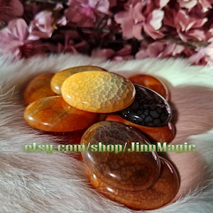 May include: A pile of polished, oval stones in shades of orange, yellow, and black. The stones have a textured, patterned surface. The Etsy shop name "JinnMagic" is visible in the image.