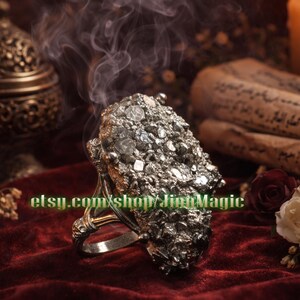 Op de afbeelding: Een grote, sierlijke ring met een getextureerde, zilverkleurige stenen zetting. De ring is gezet op een goudkleurige band en rust op een dieprode fluwelen ondergrond. De Etsy shopnaam "etsy.com/shop/JinhMagic" is zichtbaar.