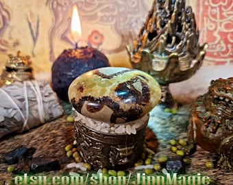 May include: A polished, oval-shaped stone with brown and green patterns sits atop a small, ornate metal container filled with white pebbles. A lit candle, a brass incense holder, and other decorative items are in the background. The Etsy shop URL is visible at the bottom.