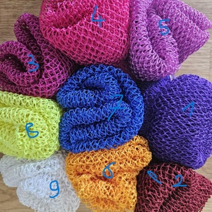 Peut inclure: Neuf poufs de bain en maille de différentes couleurs, dont rose, violet, bleu, jaune, blanc, orange et marron. Les poufs sont disposés en cercle sur une surface en bois.