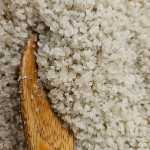 Puede incluir: Primer plano de un montón de sal marina gruesa y blanca con una cuchara de madera parcialmente sumergida en la sal.