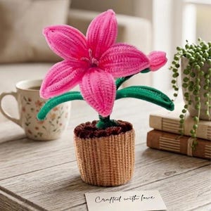 May include: A handcrafted pink flower made from a fuzzy material, with green stem and leaves, in a woven basket pot. The flower is on a wooden table with a coffee mug and a card that says "Crafted with love".