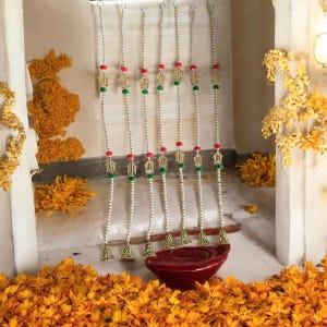 Puede incluir: Adornos colgantes decorativos con cuentas con detalles dorados, cuentas rojas y verdes y pequeñas campanas doradas. Los adornos cuelgan frente a un cuenco rojo oscuro, rodeado de flores naranjas.