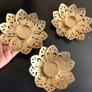 May include: Three gold-coloured, flower-shaped candle holders. Each holder has a central circular space for a candle and petal-shaped cutouts. The holders are made of metal and have a matte finish.