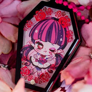 May include: A black coffin-shaped charm with a cartoon girl with pink and purple hair, framed by red roses. The charm hangs on a pink beaded chain, set against a backdrop of pink and purple flowers.