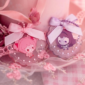 May include: Two circular acrylic charms featuring cartoon characters, one in shades of pink and one in shades of purple. Each charm has a scalloped edge, a small ball chain, and a satin ribbon bow attached at the top, displaying a cute illustration.
