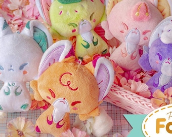 Puede incluir: Cinco juguetes de peluche de zorro en una cesta con flores rosas. Los zorros son de diferentes colores, incluyendo blanco, verde, rosa, naranja y morado. Cada zorro tiene un diseño diferente en su cuerpo. El texto "Petit Foxies Collection" es visible en la esquina inferior derecha de la imagen.