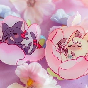 May include: Two adorable cartoon animals sleeping inside pink flower-shaped charms. The cat is purple and the dog is white with a pink bow.