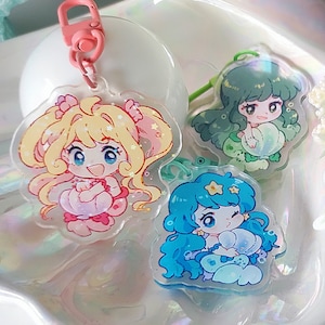 May include: Three colourful character charms featuring a blonde girl with a pink dress, a green-haired girl with a blue dress, and a blue-haired girl with a blue dress. The charms are made of clear plastic and have a glossy finish.