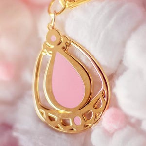 May include: A gold-colored teardrop-shaped charm with a pink center and a gold clasp. The charm has a decorative openwork design and is suspended from a gold-colored clasp. The background is a soft, blurred pink and white.