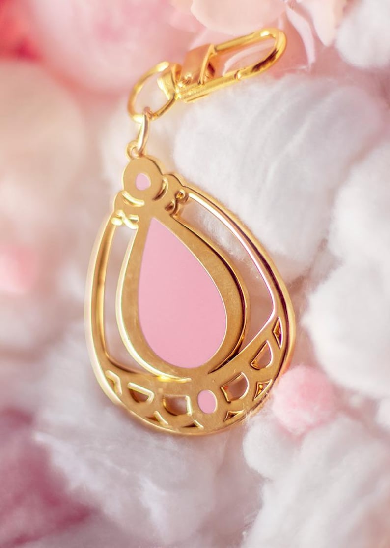 May include: A gold-colored teardrop-shaped charm with a pink center and a gold clasp. The charm has a decorative openwork design and is suspended from a gold-colored clasp. The background is a soft, blurred pink and white.