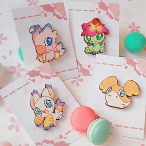 Può includere: Quattro spille in legno con personaggi dei cartoni animati della serie anime Digimon. Le spille sono su carte bianche con disegni floreali rosa. I personaggi sono un uccello rosa, un mostro vegetale verde, una creatura felina bianca e gialla e una creatura canina gialla.