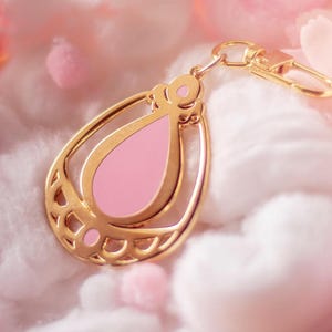 May include: A gold-colored teardrop-shaped keychain with a pink enamel center. The keychain has a decorative design and a clasp for attaching to keys or a bag. The background is a soft, blurred pink and white.