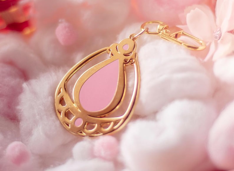 May include: A gold-colored teardrop-shaped keychain with a pink enamel center. The keychain has a decorative design and a clasp for attaching to keys or a bag. The background is a soft, blurred pink and white.