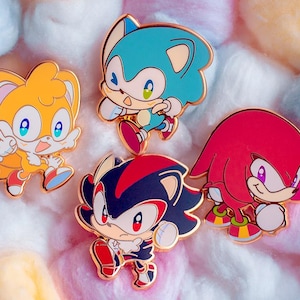 Op de afbeelding: Vier emaillepins met personages uit de Sonic the Hedgehog-videogameserie. De pins zijn van Sonic, Tails, Shadow en Knuckles. De pins bevinden zich op een roze en witte achtergrond.