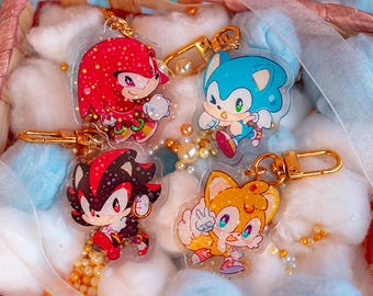 May include: Four colorful character keychains featuring Sonic the Hedgehog, Knuckles the Echidna, Shadow the Hedgehog, and Tails the Fox. The keychains are made of clear plastic with glitter embedded in the plastic. The characters are depicted in a cartoon style.