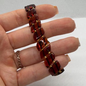 K&ouml;nnte beinhalten: Ein Vintage-Armband mit rechteckigen, bernsteinfarbenen Segmenten mit dunklen Streifen. Die Segmente sind durch kleine goldfarbene Metallst&auml;be verbunden. Das Armband hat eine warme, rotbraune Farbe und wirkt durchscheinend.