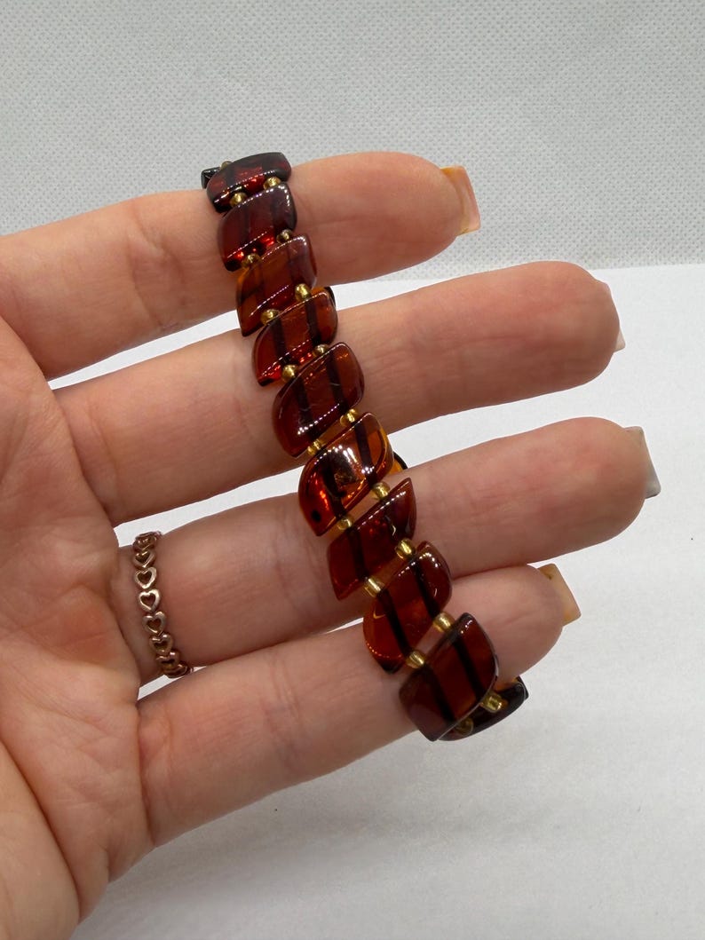 K&ouml;nnte beinhalten: Ein Vintage-Armband mit rechteckigen, bernsteinfarbenen Segmenten mit dunklen Streifen. Die Segmente sind durch kleine goldfarbene Metallst&auml;be verbunden. Das Armband hat eine warme, rotbraune Farbe und wirkt durchscheinend.