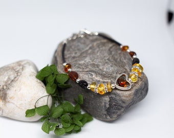 Könnte beinhalten: Ein silbernes Herz-Charm-Armband mit Bernsteinperlen in Gelb-, Orange- und Brauntönen. Das Armband ist auf einem schwarzen Band und wird auf einem grauen Stein präsentiert.