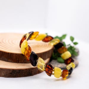 K&ouml;nnte beinhalten: Ein mehrfarbiges Bernsteinarmband mit polierten Steinen in Gelb-, Orange- und Braunt&ouml;nen, verbunden durch goldfarbene Glieder. Die Bernsteinst&uuml;cke sind unregelm&auml;&szlig;ig geformt. Das Armband wird auf einer Holzoberfl&auml;che pr&auml;sentiert.