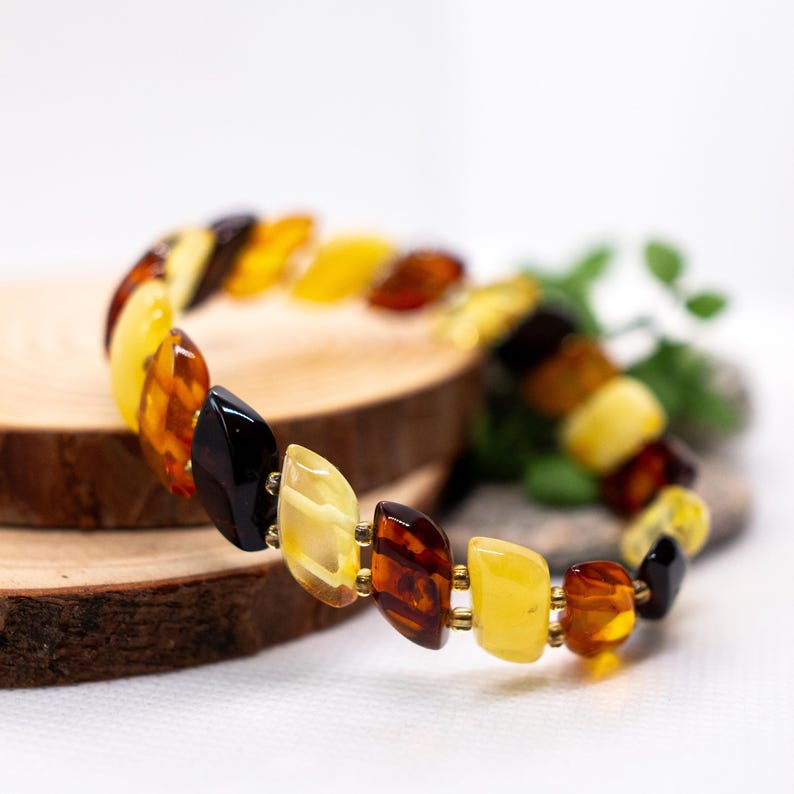 K&ouml;nnte beinhalten: Ein mehrfarbiges Bernsteinarmband mit polierten Steinen in Gelb-, Braun- und Schwarzt&ouml;nen. Die Bernstein-St&uuml;cke sind in verschiedenen Formen gehalten und mit goldfarbenem Metall verbunden. Das Armband liegt auf einer Holzoberfl&auml;che.