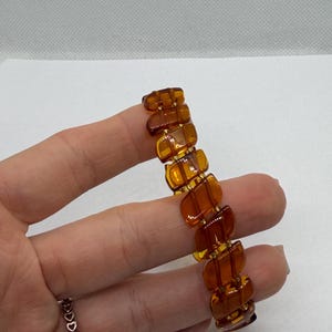 K&ouml;nnte beinhalten: Ein Vintage-Armband in Bernsteinfarbe mit einem sich wiederholenden rechteckigen Design. Das Armband besteht aus durchscheinenden, polierten Segmenten, die mit goldfarbenem Metall verbunden sind. Das Armband wird in einer Hand gehalten und zeigt seine warmen, honigartigen T&ouml;ne.