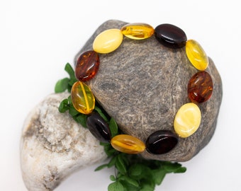 Könnte beinhalten: Ein mehrfarbiges Bernsteinarmband mit ovalen Perlen in Gelb-, Braun- und Schwarztönen. Das Armband liegt auf einem grauen Stein mit grünem Laub vor weißem Hintergrund. Die Perlen sind etwa 2 cm lang.