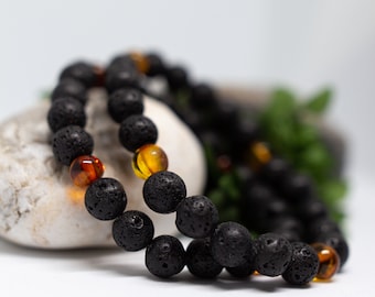 Könnte beinhalten: Ein Armband aus schwarzen Lavasteinen und Bernsteinperlen. Das Armband besteht aus kleinen, runden Perlen und wird auf einer weißen Oberfläche gezeigt.