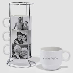 May include: A white ceramic mug set with a metal stand. The stacked mugs display black and white photos of couples. A single mug with the text "Love at first sip" is also present.