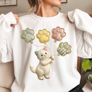 Puede incluir: Sudadera blanca con un gatito cosido que sostiene globos con forma de pata. Los globos son de colores pastel con estampados florales y de cuadros. El gatito tiene un lazo rosa y sostiene los globos con una cuerda.