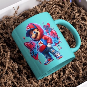 May include: A teal ceramic mug featuring a cartoon illustration of a character in a red hoodie and blue jeans. The design includes graffiti-style lettering and stars. The mug is presented in a white box with brown paper filler.
